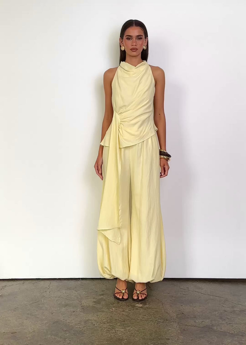 Video: Capriana Set in Light Yellow with a draped high neckline, sleeveless top, gathered side waist with a long cascading sash, and relaxed wide-leg pants with a softly gathered ankle-length hem.