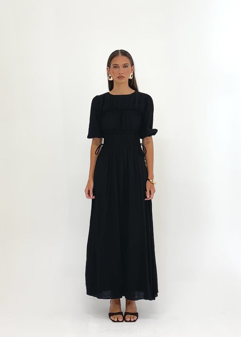 Video: Nadinia Maxi Dress in Black, featuring a round neckline, softly gathered bodice, short puff sleeves with elasticated cuffs, a fitted panelled waist with adjustable side ties, and a flowing maxi-length silhouette.