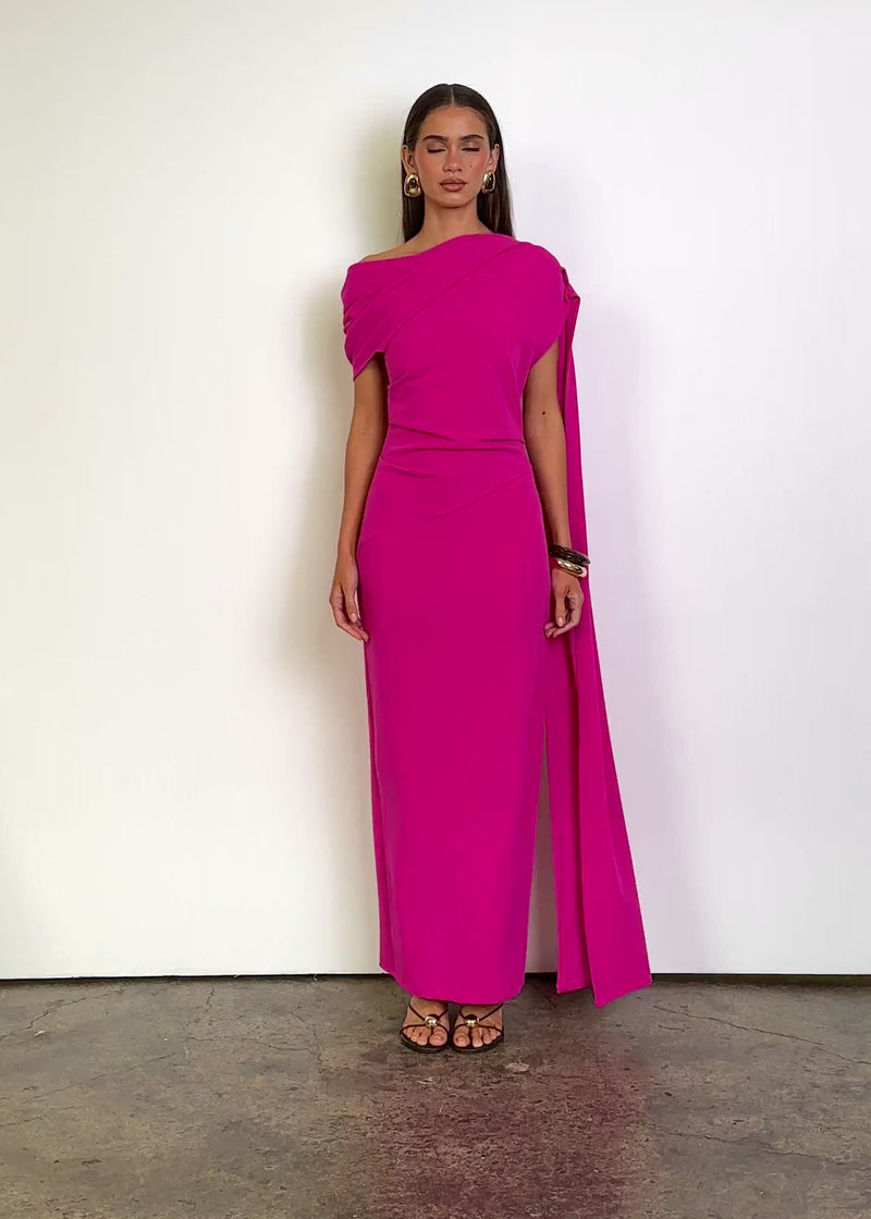 Video: Jovania Maxi Dress in Hot Pink with an asymmetric draped neckline, cap-style shoulder coverage, fitted silhouette, diagonal waist seam and a floor-length hem, finished with a long cascading shoulder sash detail.