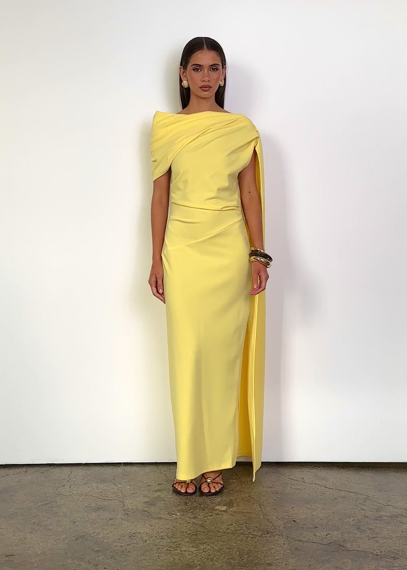 Video: Jovania Maxi Dress in Lemon with a softly draped asymmetric neckline, short sleeves and a fitted silhouette, featuring layered shoulder draping, a long flowing sash detail, subtle waist shaping and a floor-length hem.