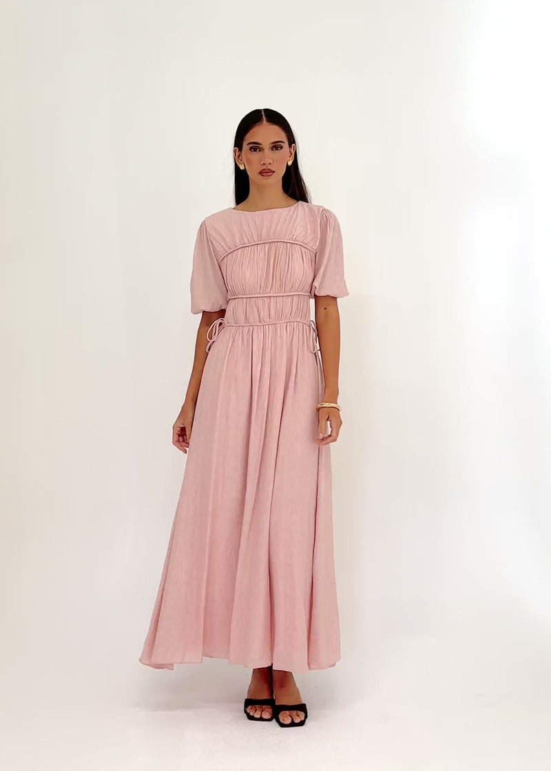 Video: Nadinia Maxi Dress in Pink, featuring a round neckline, softly gathered bodice, short puff sleeves with elasticated cuffs, a fitted panelled waist with adjustable side ties, and a flowing maxi-length silhouette.