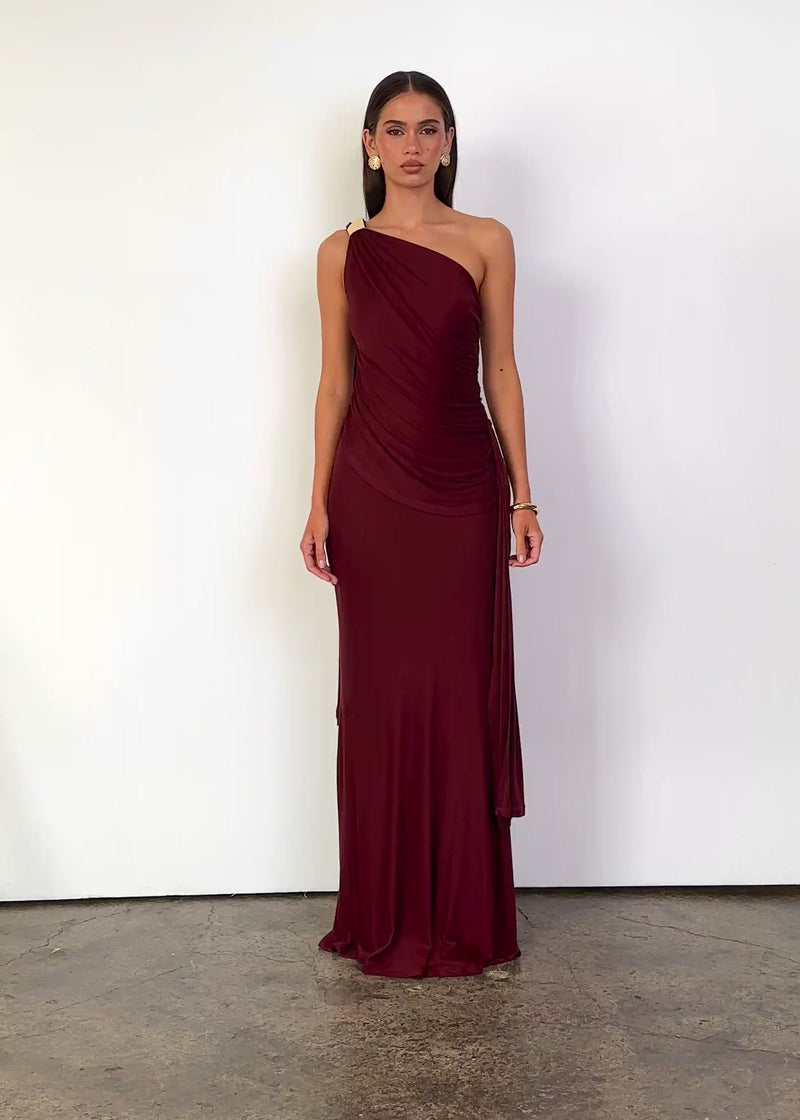 Video: Serina One Shoulder Maxi Dress in Wine with an asymmetric one-shoulder neckline, sleeveless fitted silhouette, gathered draping through the bodice and waist, gold shoulder hardware, cascading side sash detail and a floor-length hem.